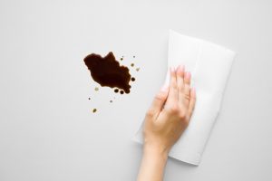 Woman's hand cleaning fresh spilled dark beverage from gray desk. Coffee stain simple removing with white paper napkin. Cleanup. Close up. Top view.